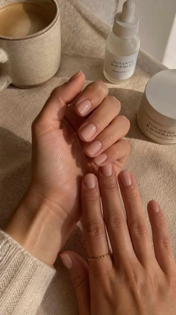 Two feminine hands displaying aesthetic minimalist natural short nails in glossy nude, blush pink, and milky neutral finishes beside skincare essentials.