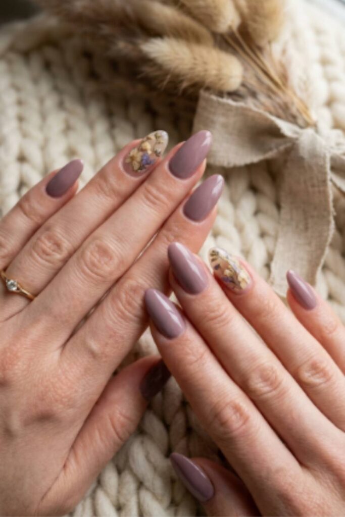 Long almond nails painted dusty mauve with delicate dried floral nail art resting on a cozy cream knit sweater.