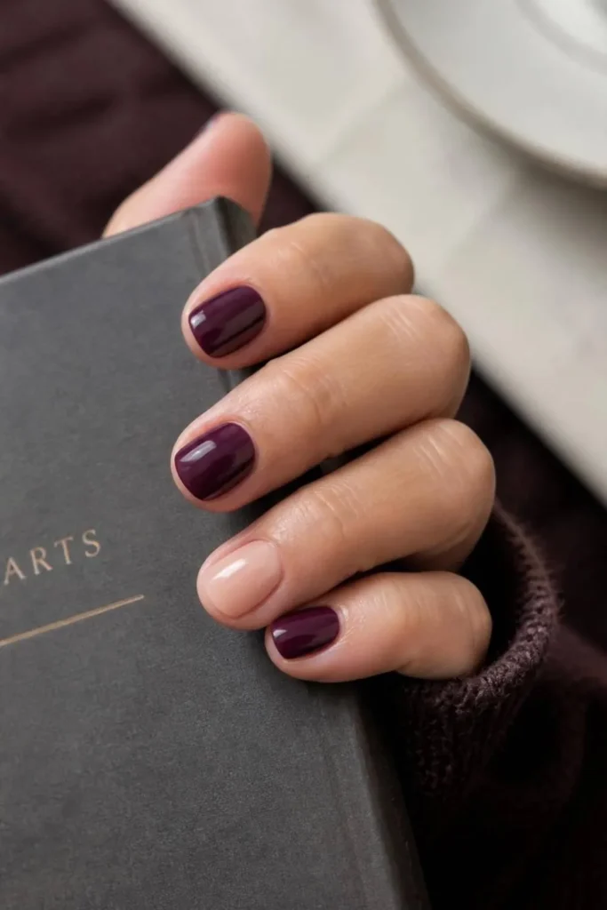 Deep plum short nails paired with one sheer glossy nude accent nail in a modern understated mismatched manicure.