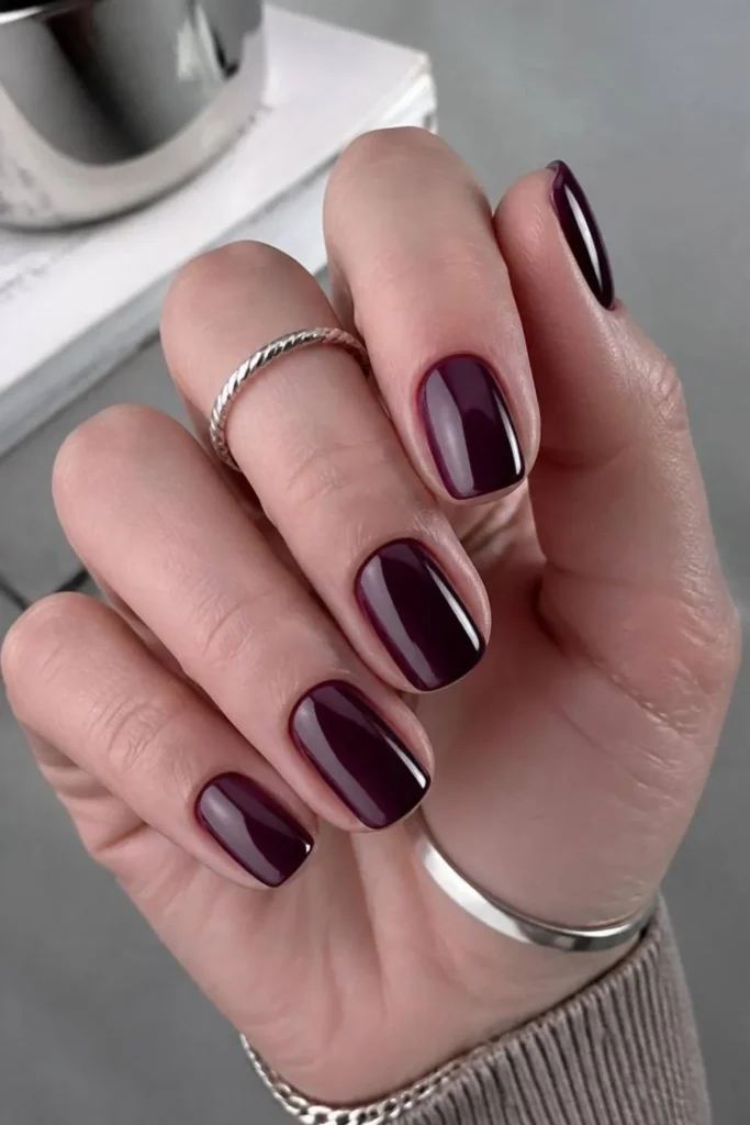 Short professional nails painted in rich plum gel polish with a glossy finish that complements cool skin undertones.