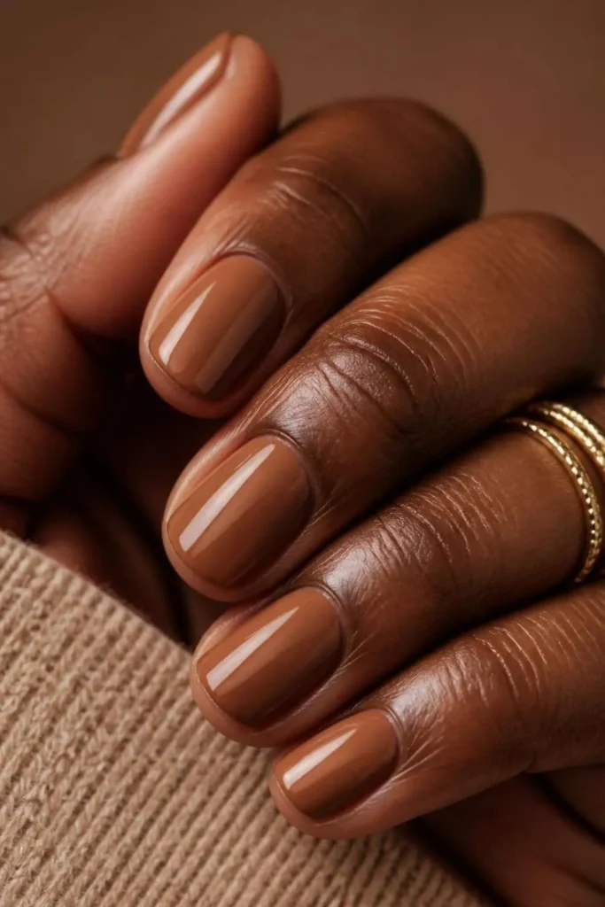 Glossy deep caramel nude polish on short natural nails complementing rich brown skin tones beautifully.