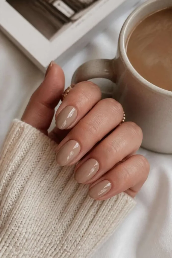 Short almond nails painted in glazed latte nude polish with ultra-glossy finish beside a ceramic coffee mug.