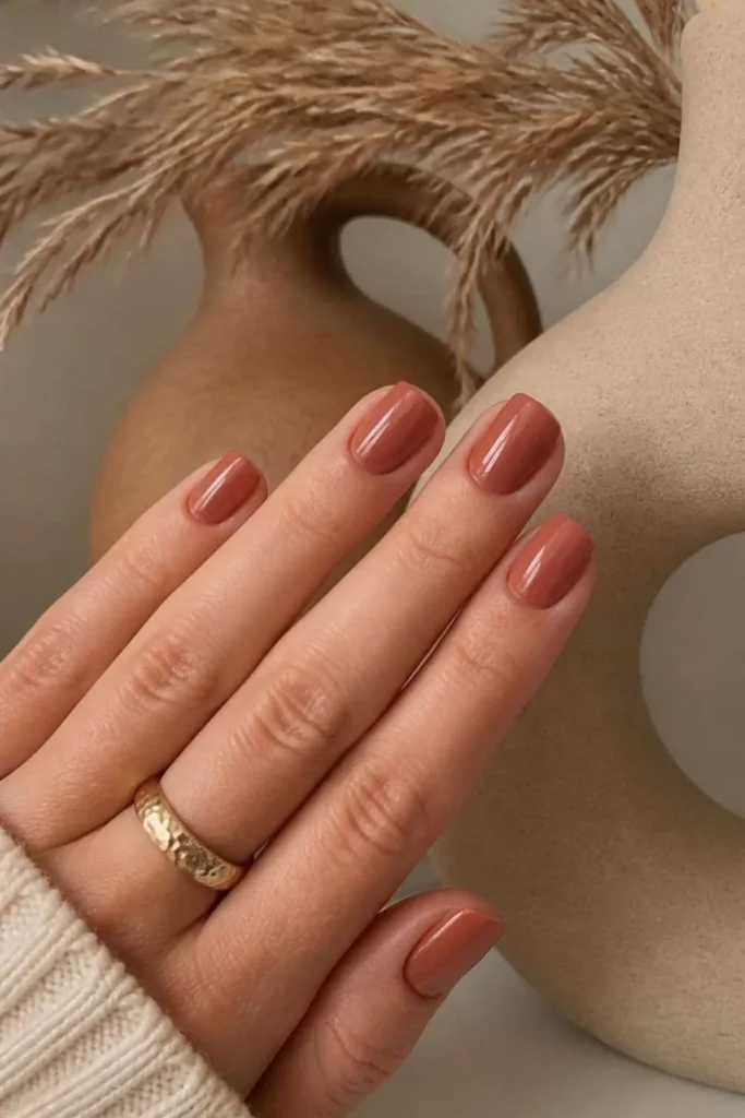 Short squoval nails painted in muted terracotta nude polish with earthy matte finish and modern styling.