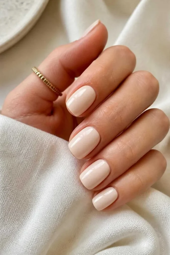 Short squoval nails painted in porcelain cream nude polish with mirror-like glossy topcoat and healthy natural finish.