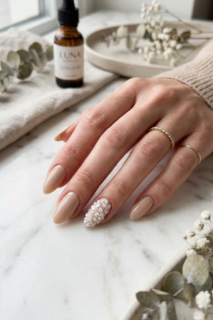 Cream almond nails with a raised embossed floral accent on the ring finger in a minimalist winter design.